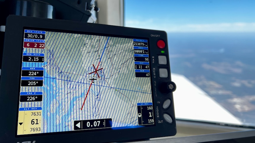 Focus on computer screen inside plane showing elevation data, with blurred view of Tweed landscape in background.