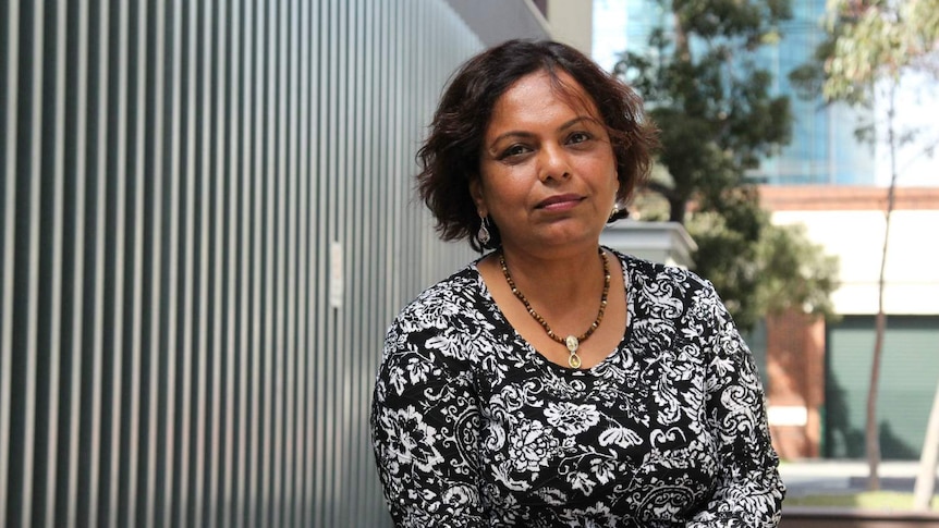 Suchitra Chari sits and looks at the camera in a laneway with grass, trees and benches.