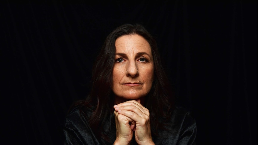 A woman with brown hair and brown eyes holding her hands underneath her chin, black background
