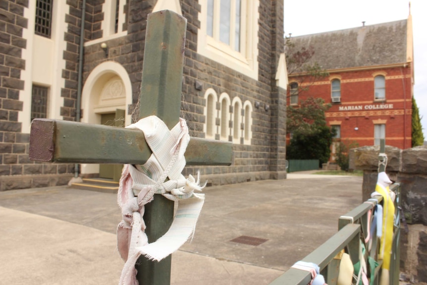 A cross with a white ribbon tied around it next to a gate also hung with ribbons outside a church with brown bricks.