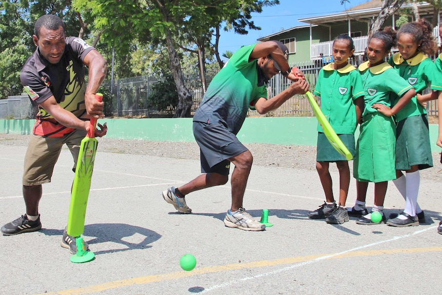 Two coaches demonstrate a forward defensive stroke to a group of girls in green school uniforms.