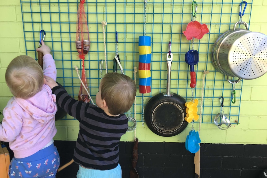 Two toddlers reach and play with various recycled implements attached to a wall, making noise and having fun