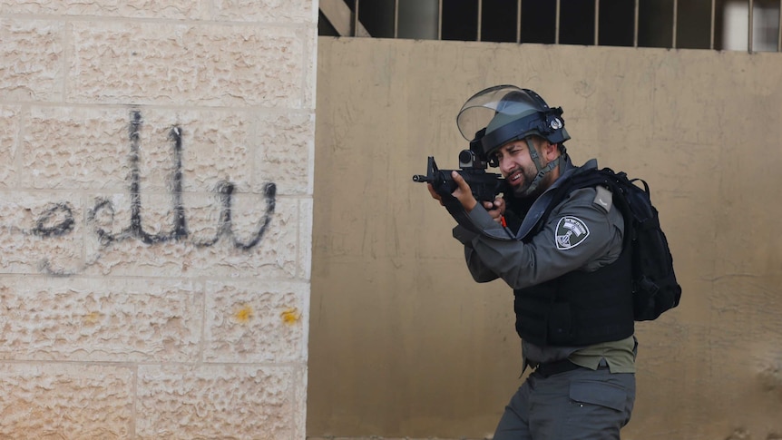An Israeli police officer peers through the viewfinder of his rifle next to a wall with graffiti in Arabic script.