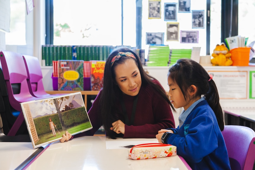An Asian Australian woman wearing a headband and maroon jumper holds a picture book up to a school aged child in a classroom