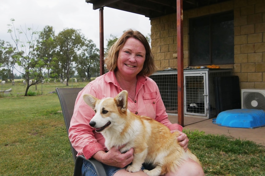 A woman in a pink shirt smiles at the camera, she is cradling a young white and fawn corgi in her lap