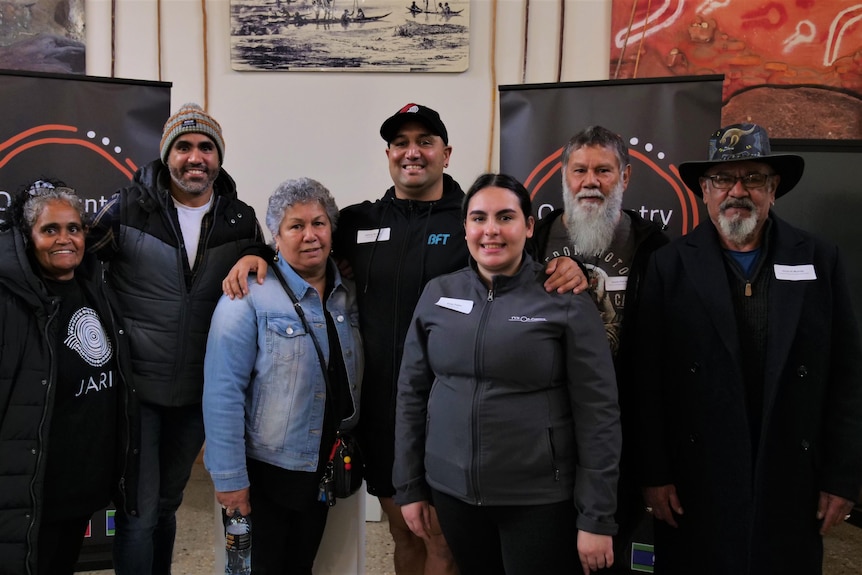 Seven people stand in front of Indigenous artwork and smile