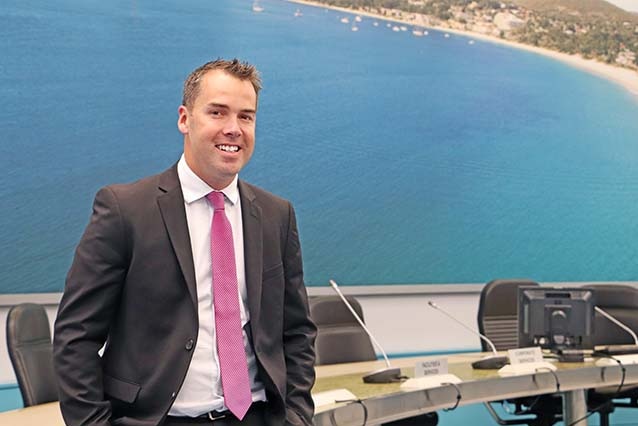 A man in a suit leans against a round council desk with a large photograph of a beach in the background.