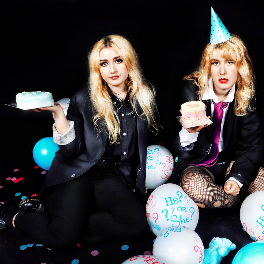 Press photo of two members of Lambrini Girls sitting in front of a black background holding cakes 