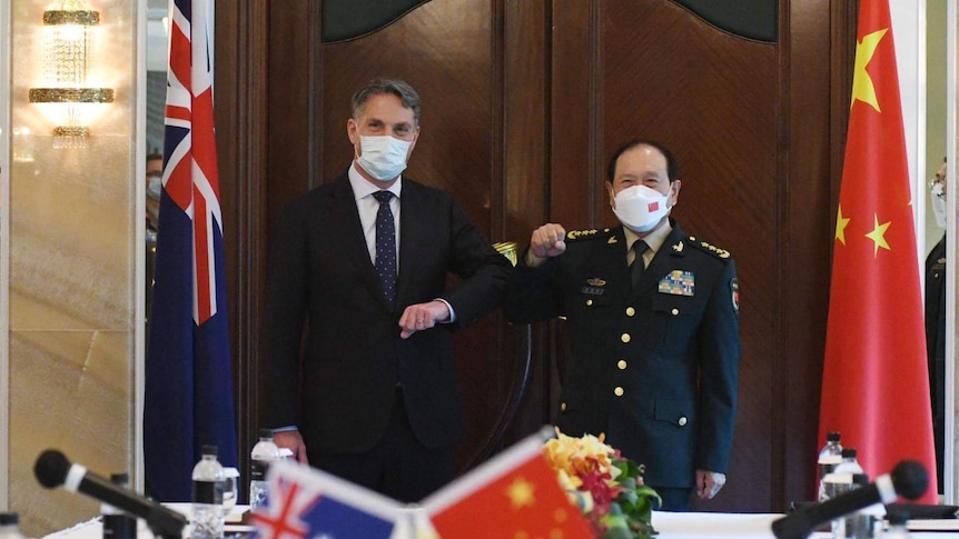 Two mask-wearing men in suits elbow bump as they pose for a photo in front of the Australian and Chinese flags.