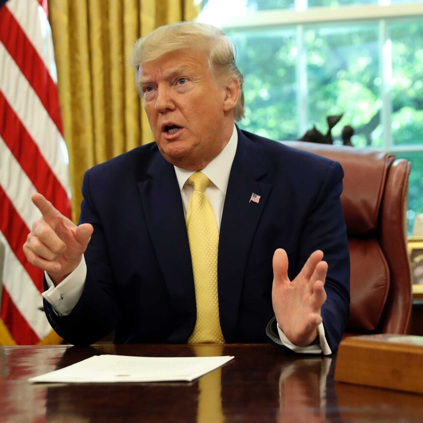 Donald Trump sits at the President's desk in the Oval Office of the White House, pointing a finger in mid-conversation.