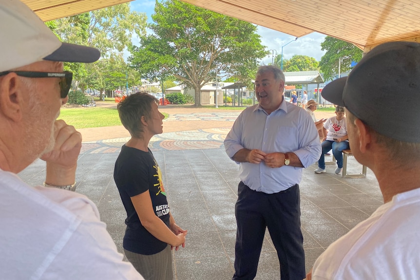 A grey-haired man speaks with some people under a shade sail in a park area.