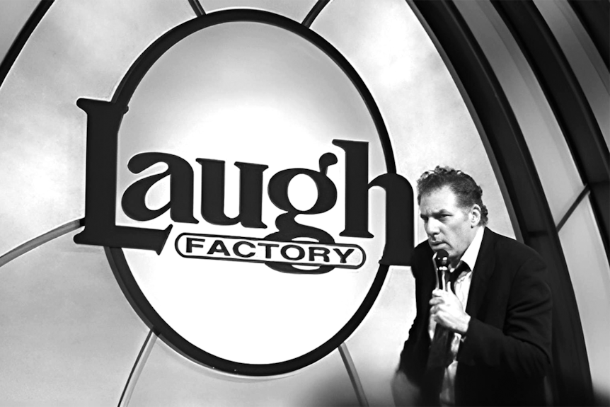 A black and white photo of Michael Richards holding a microphone standing in front of a Laugh Factory sign