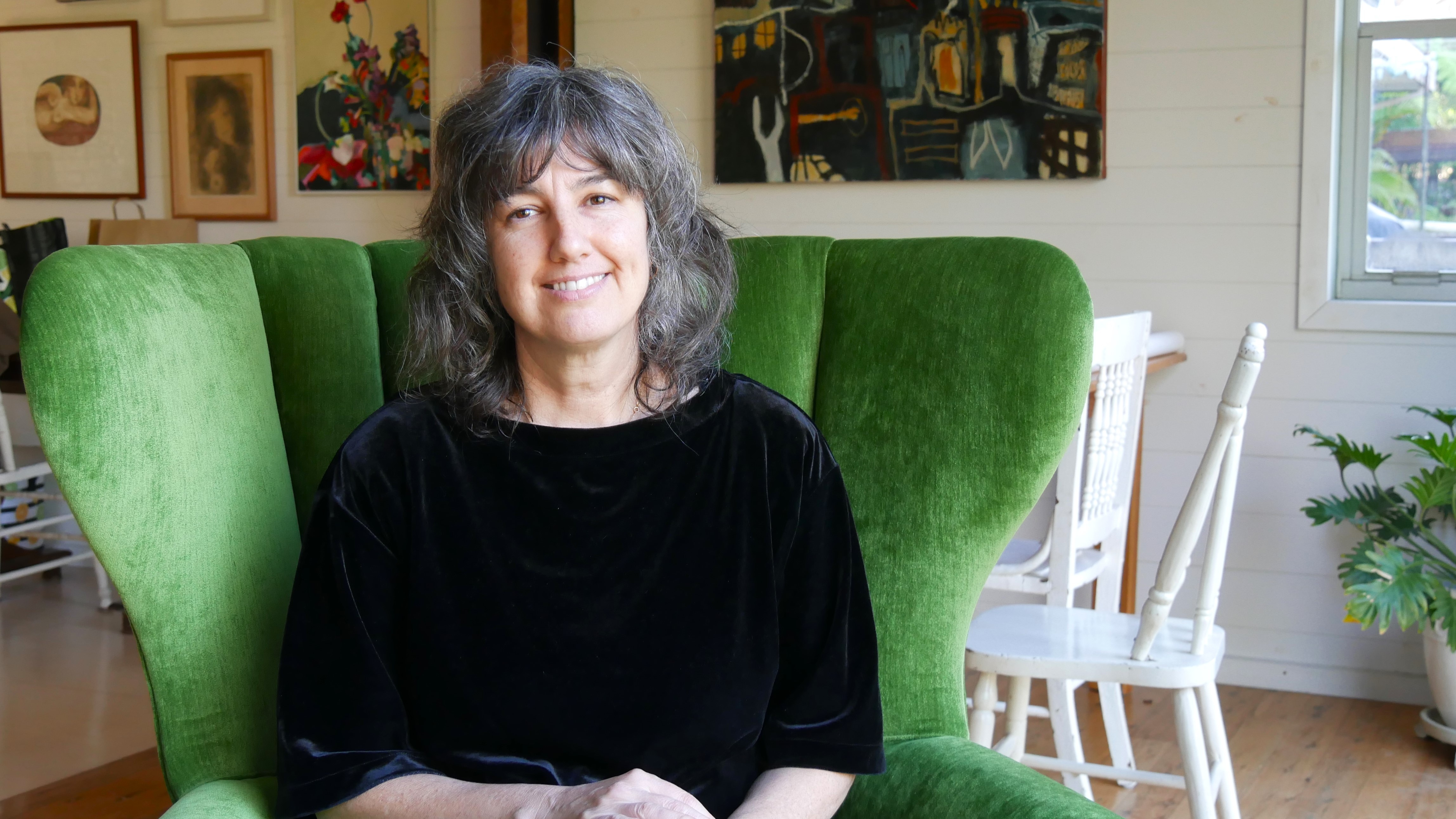 A woman wearing a black suede shirt sits in a large green chair and smiles. 