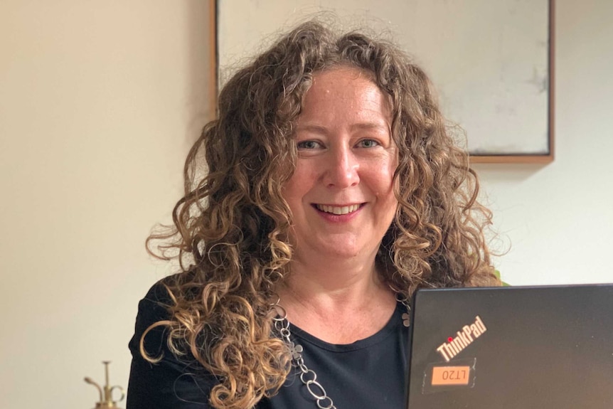 A smiling woman with long curly hair sits in front of a laptop computer.