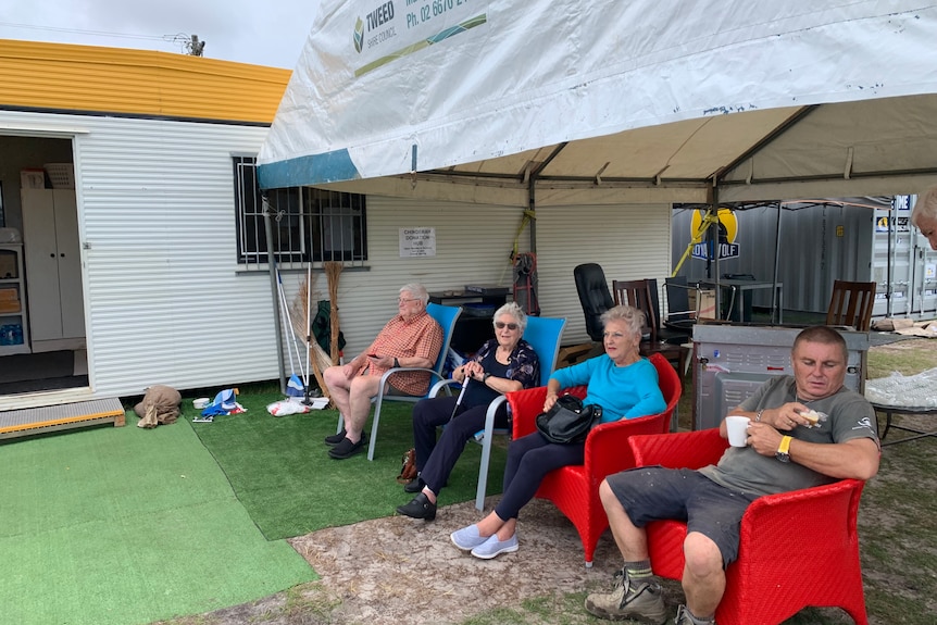 Four people sit socially distanced on chairs under an awning at a caravan park