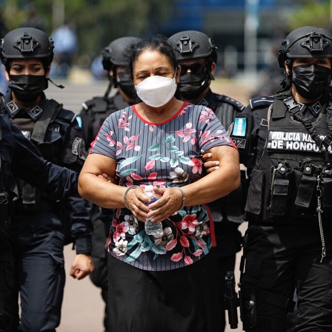 A woman wearing a mask between several highly armed policemen