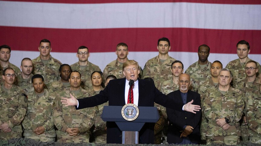 Donald Trump with arms stretched out stands at a podium in front of troops and a US flag