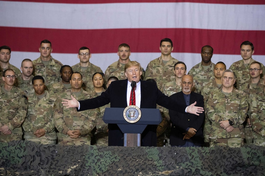 Donald Trump with arms stretched out stands at a podium in front of troops and a US flag