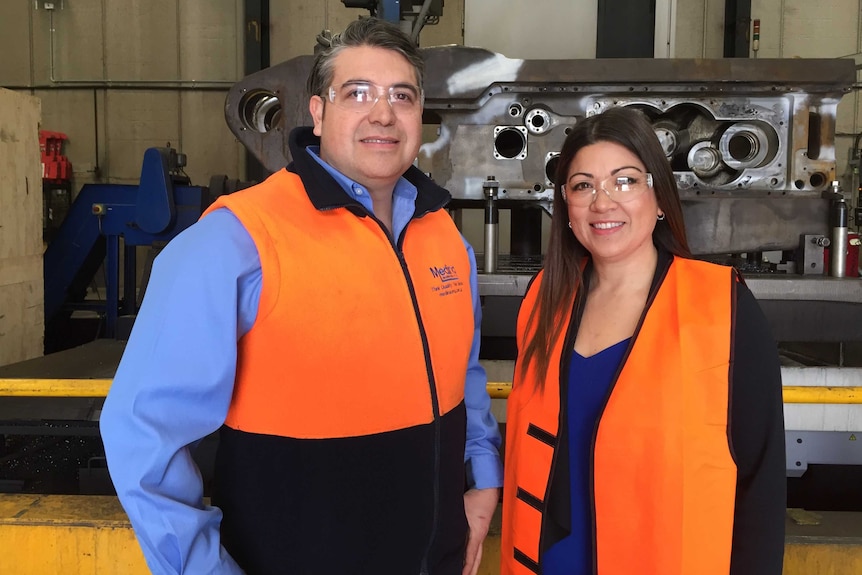 A man and a woman in high-visability vests and safety goggles standing in front of machinery.