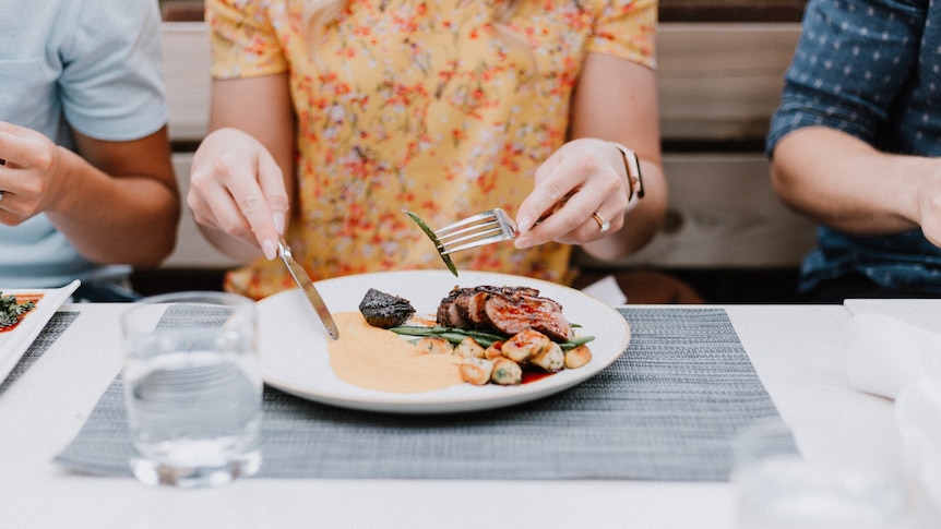 a woman sits at a table eating lunch, including red meat and potatoes.