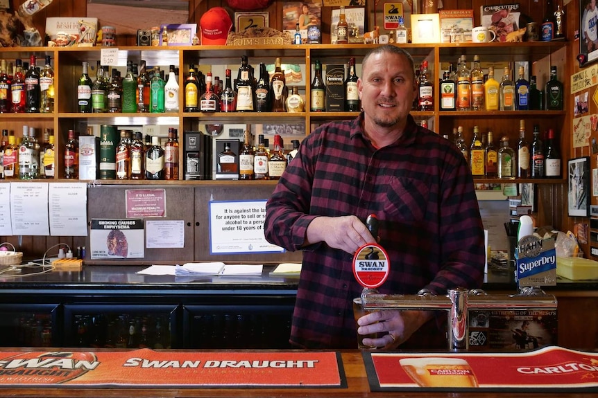 Kirk Pohl pours a beer behind the bar in the Perenjori Hotel. Shelves behind him are filled with spirit bottles and bric-a-brac.