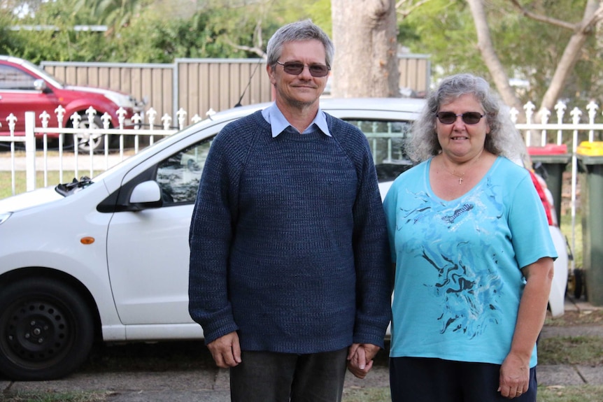 A man and a woman standing in front of their white small car, with fences, trees and a neighbour's car in background.