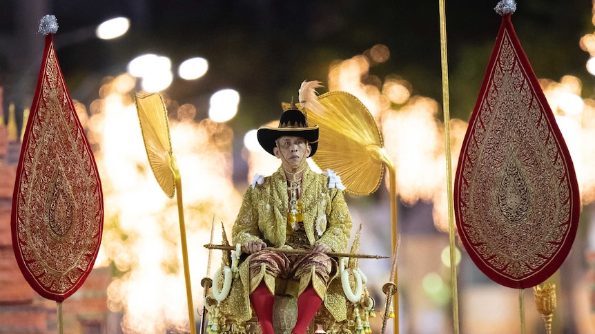 Thailand's King Maha Vajiralongkorn sits on a grand golden palanquin as fireworks explode in the distance behind him.