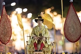 Thailand's King Maha Vajiralongkorn sits on a grand golden palanquin as fireworks explode in the distance behind him.