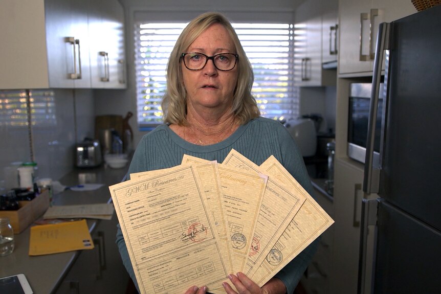 A woman holds up five share certificates, frowning, as she stands in a kitchen.