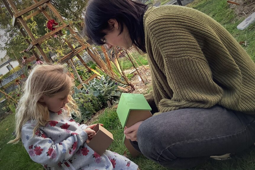 Young girl is sitting with woman in a garden, both looking into a small box that the girl is holding.