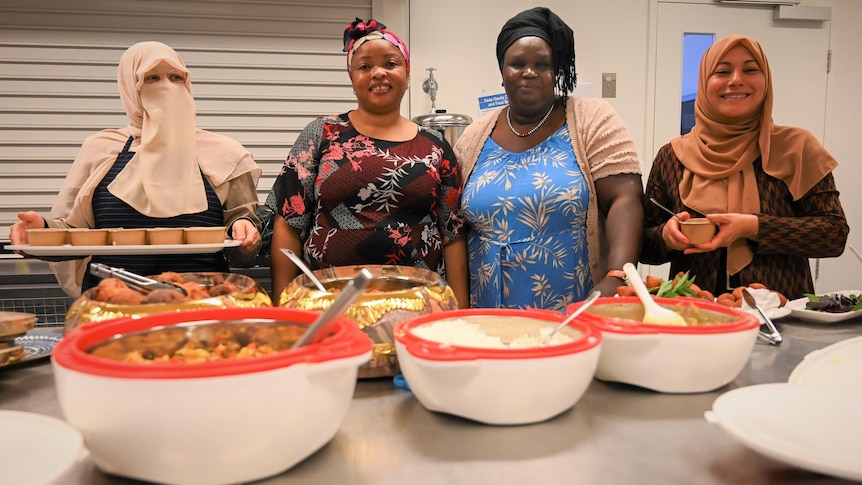 Four women stand in a row, in front of table laden with bowls of food