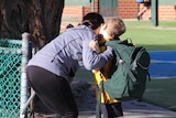 A mother in a blue top kisses her son in school uniform at the gate of a primary school while dropping him off.