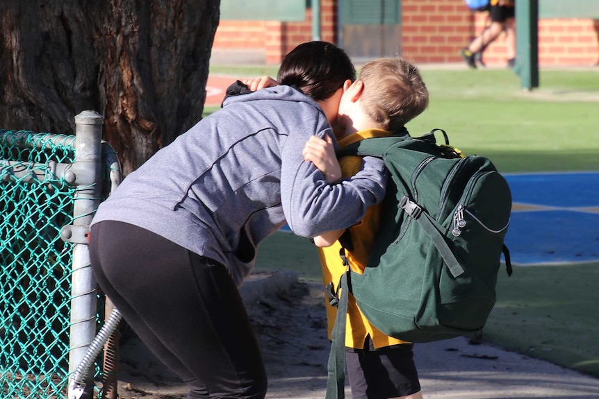 A mother in a blue top kisses her son in school uniform at the gate of a primary school while dropping him off.