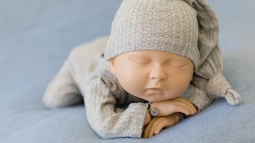 A baby mannequin in a grey jumpsuit and beanie laying belly down on a blue blanket with its arms crossed.