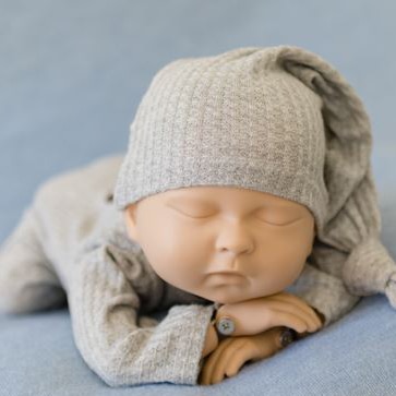 A baby mannequin in a grey jumpsuit and beanie laying belly down on a blue blanket with its arms crossed.