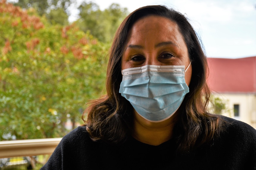 A woman with long brown air and a face mask smiles at the camera.