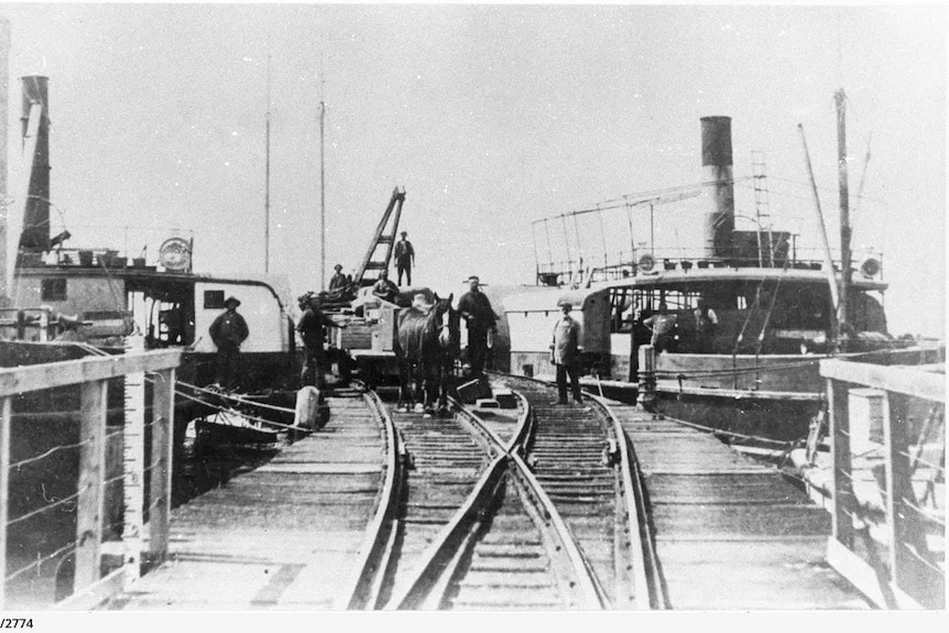 A black and white historic image of a jetty with a paddlesteamer moored on each side with a horse pulling a load on tracks