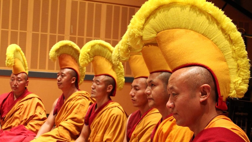 Six monks dressed in red and orange robes sitting crossed legged in a recording studio