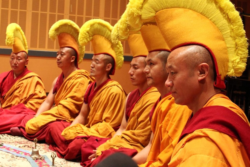 Six monks dressed in red and orange robes sitting crossed legged in a recording studio