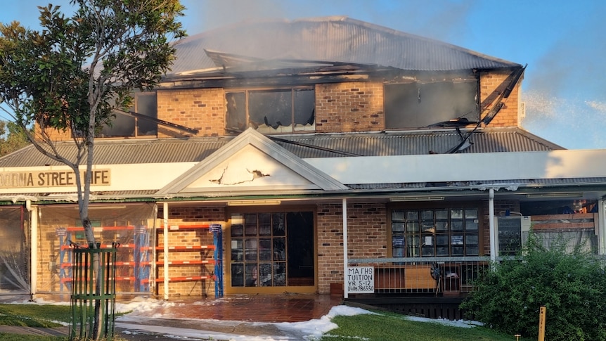 A photo of a burnt out brick building with smoke still coming out of smashed windows. It has a sign that says Goodna Street Life