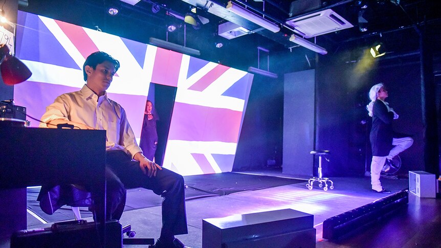 A man sits to right of stage, a woman stands in the middle of projected Union Jack flag, man in colonial wig marches.