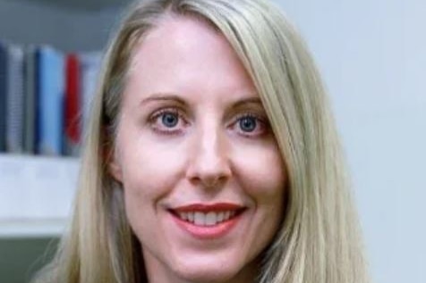 A woman with long blonde hair and blue jumper, smiles widely. A blurred bookshelf is in the background.