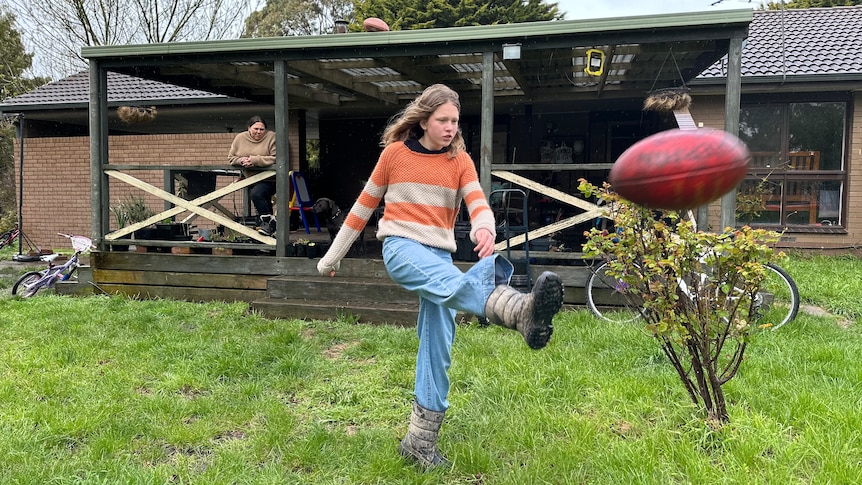 A girl kicking a football in a backyard, while an adult watches on from the verandah.