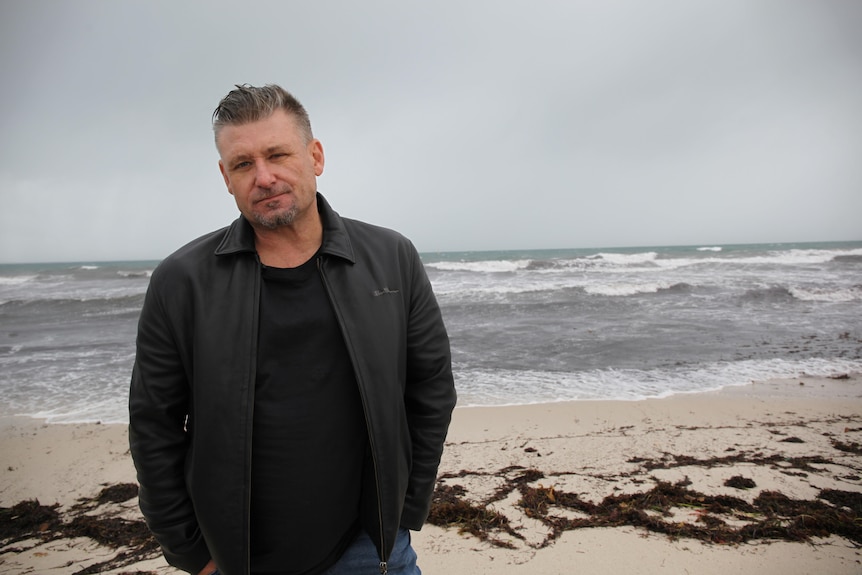 A middle-aged man wearing dark clothes stands with the ocean and grey skies behind him