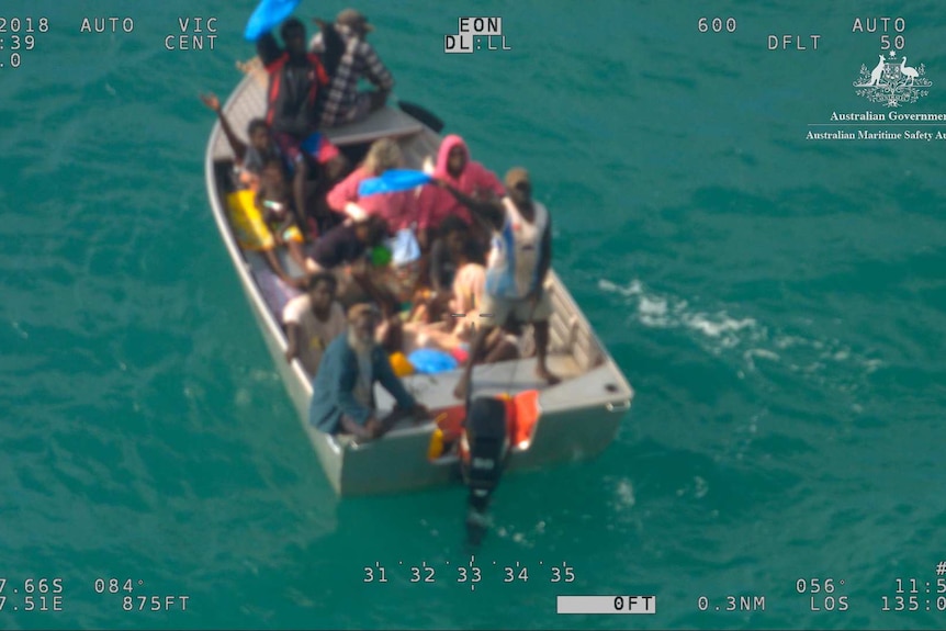A small dinghy floating in the ocean with 15 people on board, some of whom are waving.