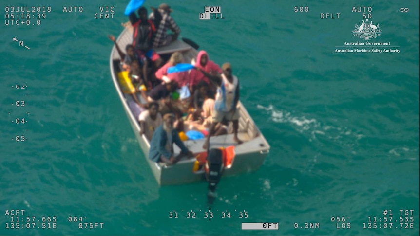 A small dinghy floating in the ocean with 15 people on board, some of whom are waving.