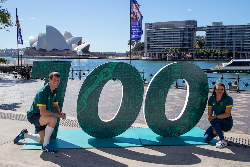 Olympians Edward Fernon and Jess Fox pose on either side of a 100 sign in front of Sydney Harbour.