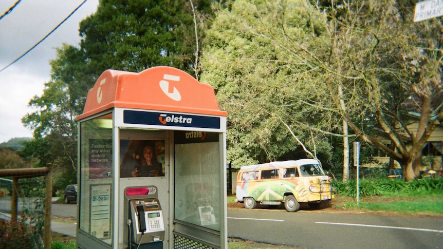 A Telstra payphone booth in foreground, a suburban leafy street in background with a VW hippie van parked in background.