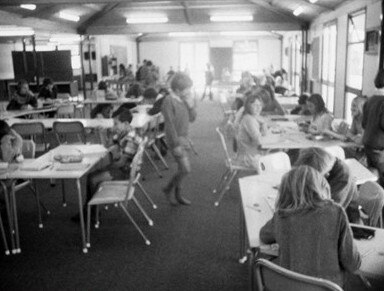 Black and white photo of children sitting at desks in a large, open classroom.