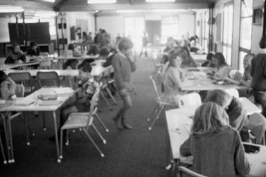 Black and white photo of children sitting at desks in a large, open classroom.
